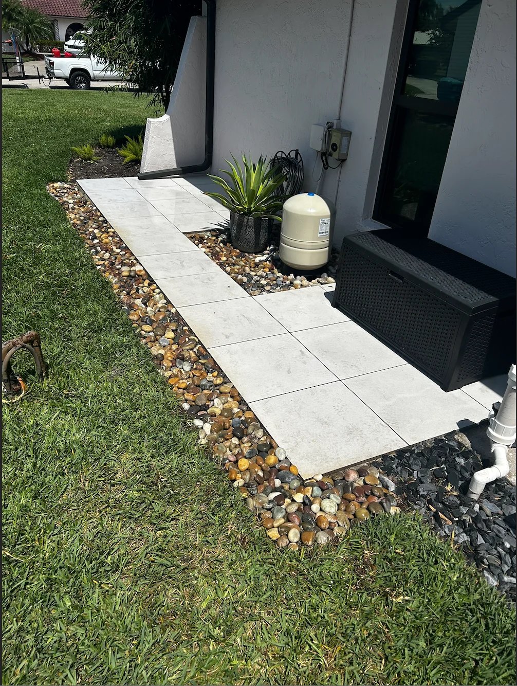 Modern patio entrance with white tile walkway bordered by decorative stones, potted plants, and white cylindrical water tank next to black door