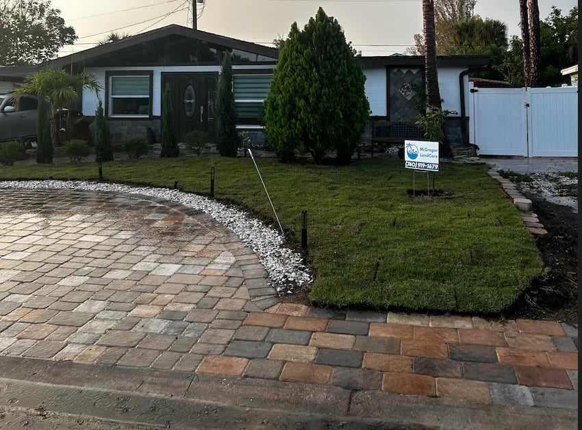 Single-story house with black roof, white and dark siding, landscaped front yard with brick patio, manicured lawn, and white gate
