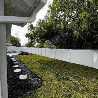 Backyard with white fence, freshly seeded lawn, black mulch strip with stepping stones, and mature trees in background
