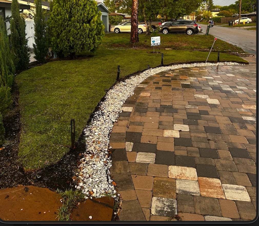Residential driveway with brown and tan pavers bordered by white gravel and green lawn, with parked cars visible in background